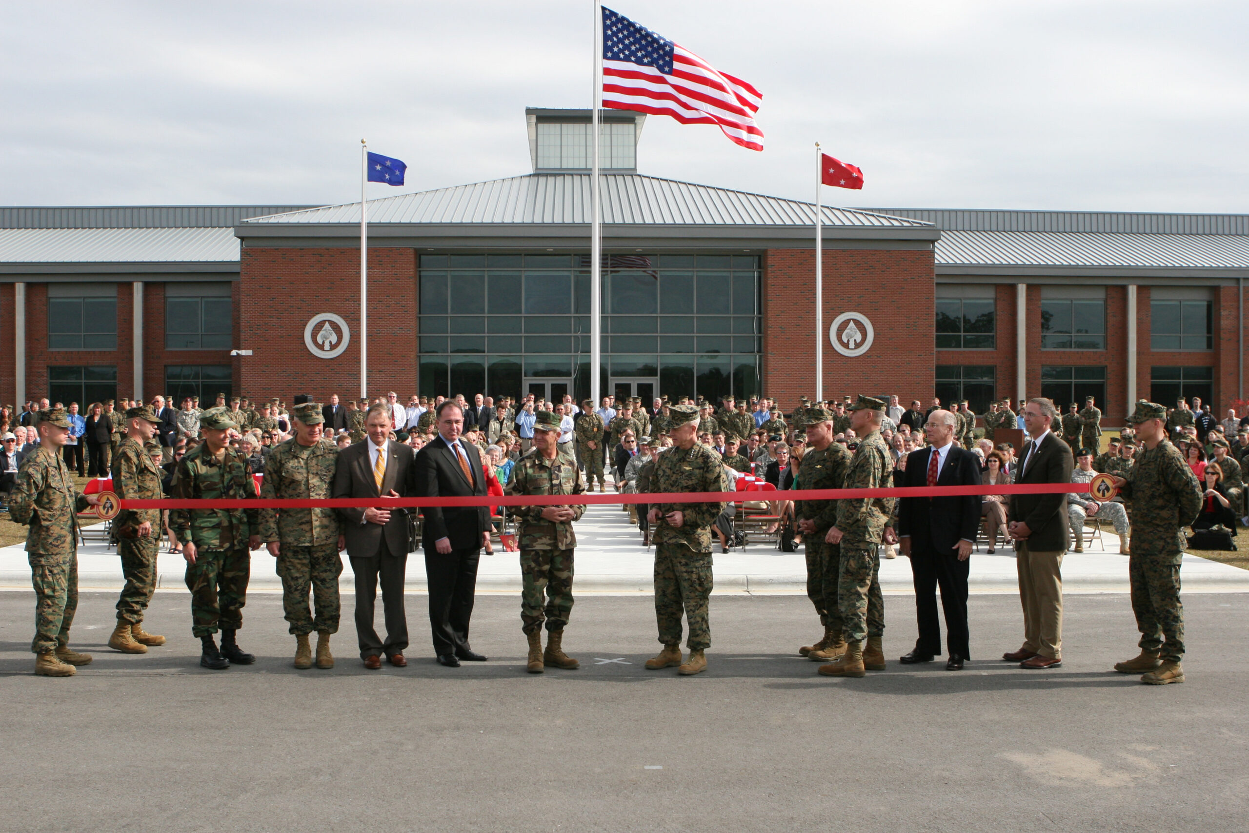 Military and civilians hold red ribbon at building opening ceremony.