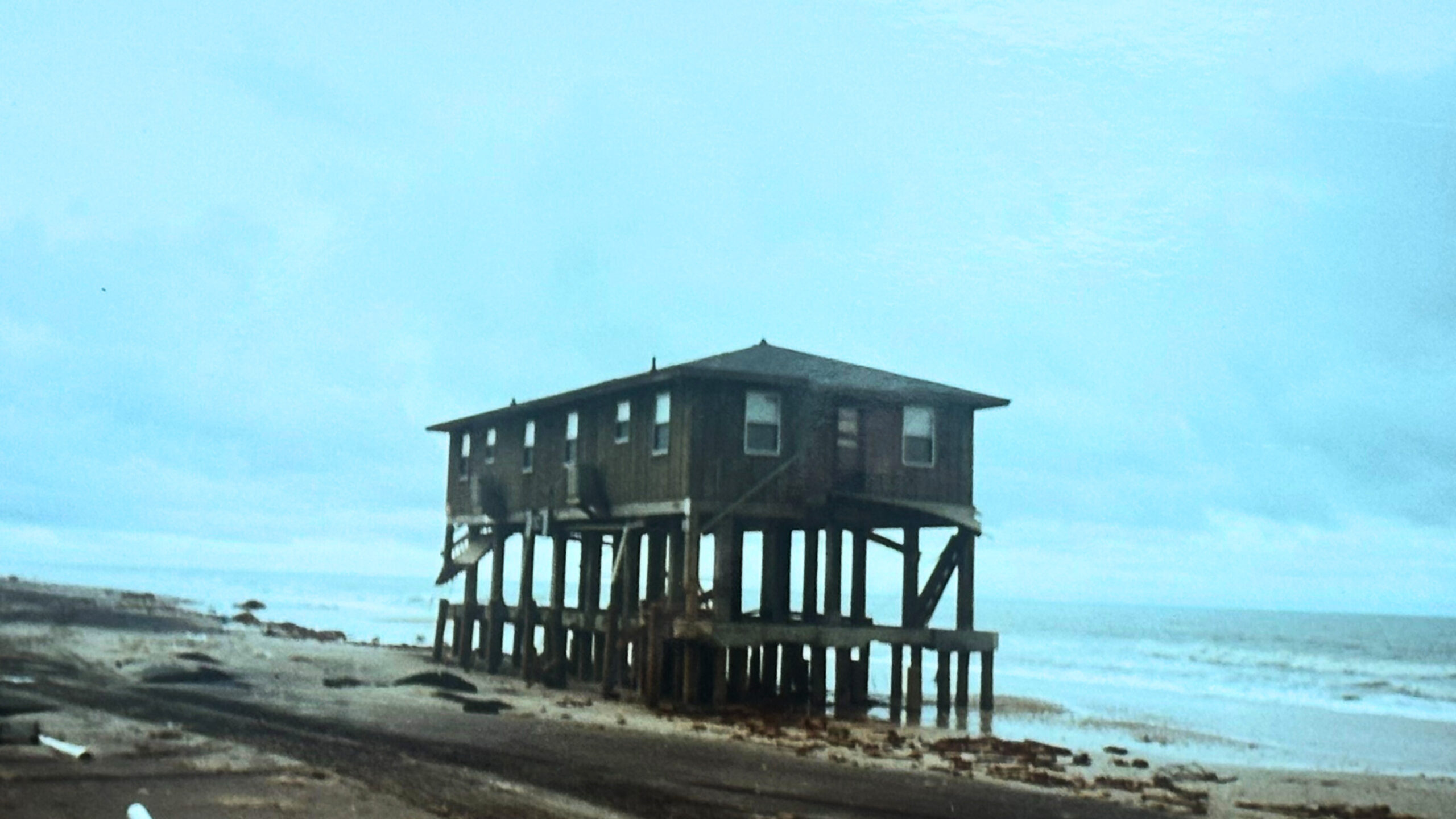 Weathered wooden house on stilts overlooks sandy beach and ocean.