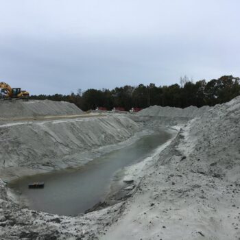 Excavator beside water-filled pit, dirt piles, cloudy sky.