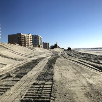Beachfront buildings, sandy beach, tire tracks, construction gear, blue sky.