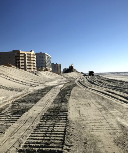Beachfront buildings, sandy beach, tire tracks, construction gear, blue sky.