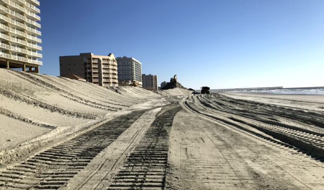 Beachfront buildings, sandy beach, tire tracks, construction gear, blue sky.