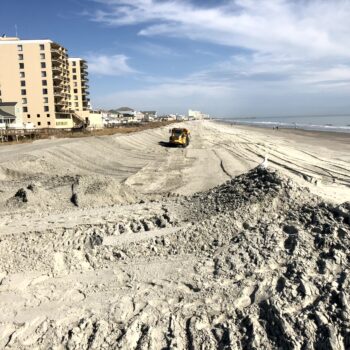 Bulldozer moves sand on beach near oceanfront buildings.